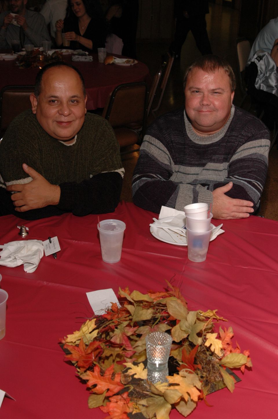 Two men sit at a red-clothed table with a fall-themed centerpiece