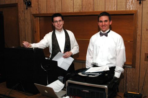 1985 Ford Crown Victoria LTD - Two men in formal attire are positioned behind a DJ setup, with a laptop and audio equipment visible.