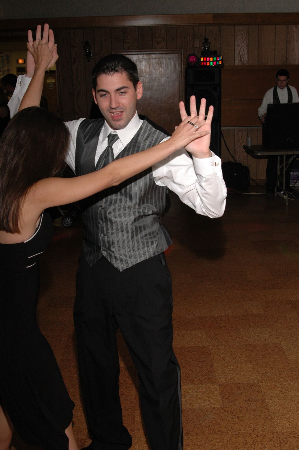Man in vest and tie dancing with a woman in a black dress at a formal event with a DJ booth in the background