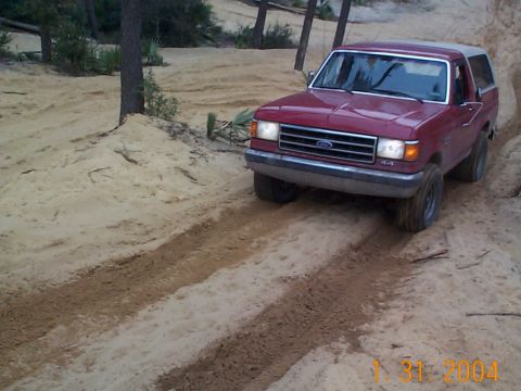 1992 Ford Bronco - A red Ford Bronco navigates a sandy off-road path with trees in the background.