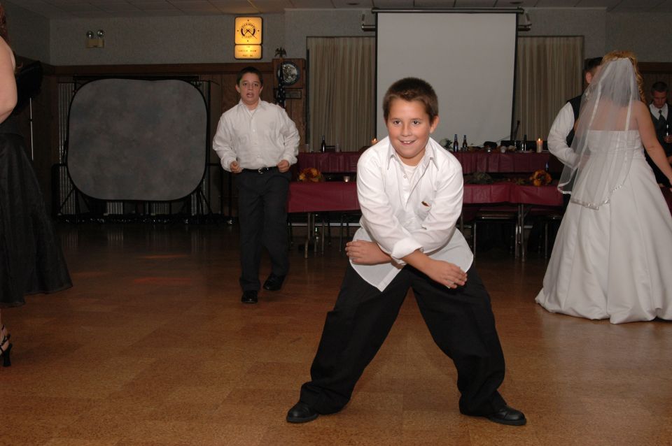 Wedding reception with two boys in white shirts and black pants, one in foreground posing, bride in background