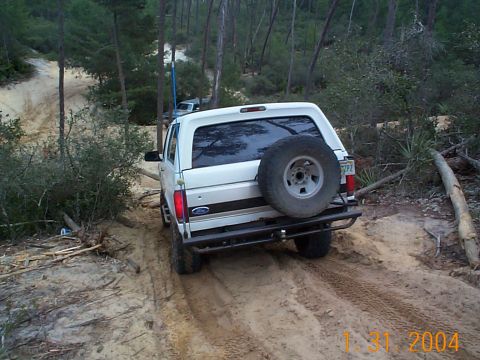 1992 Ford Bronco - A white Ford Bronco navigates a sandy, wooded trail, kicking up dust as it goes.