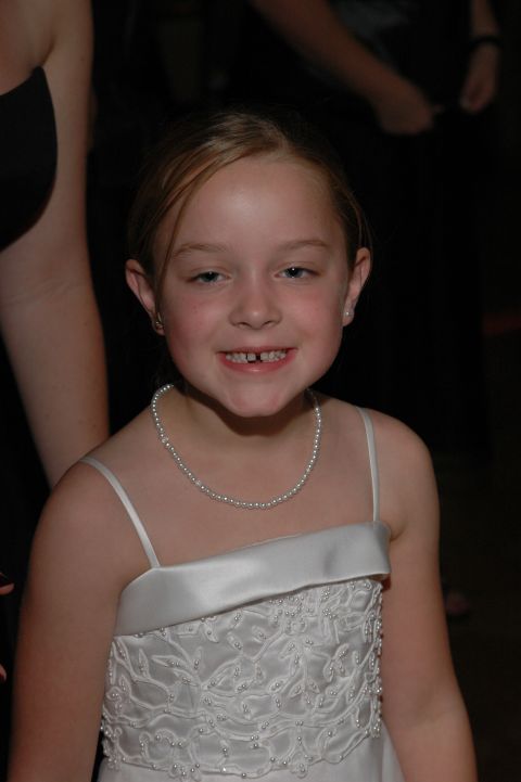 Young girl in a white dress and pearl necklace smiles, showing missing teeth
