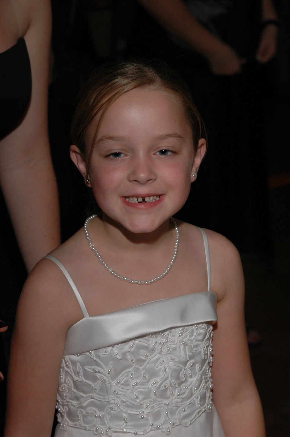 Young girl in a white dress and pearl necklace smiles, showing missing teeth