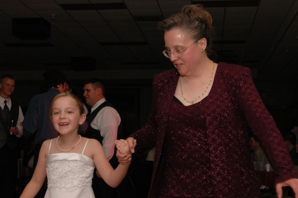 Woman in a sparkly maroon jacket and a young girl in a white dress hold hands while dancing
