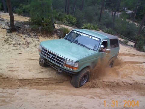 1992 Ford Bronco - A green 1992 Ford Bronco navigates a sandy off-road trail, kicking up dirt as it goes.