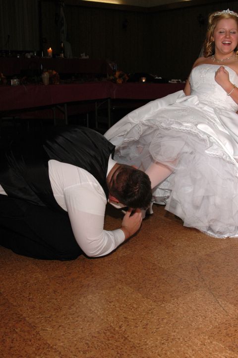 Groom in black vest and white shirt kneels to kiss bride's shoe, bride in white wedding dress laughs