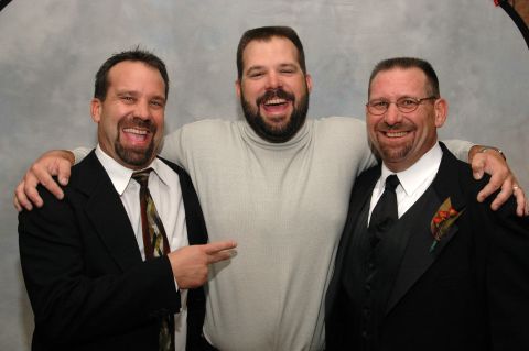 Three men in suits and a turtleneck smile for a photo against a gray backdrop