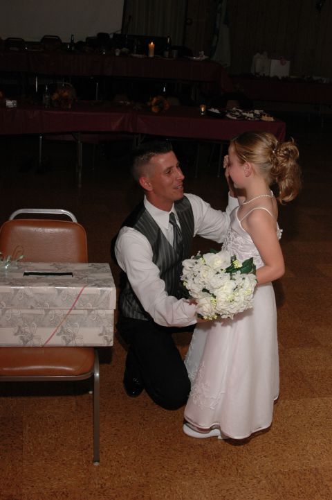 Man in a vest and tie kneels before a young girl in a white dress holding a bouquet
