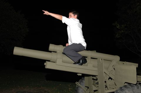 Person in white shirt and dark pants sits on a tan military howitzer, pointing left
