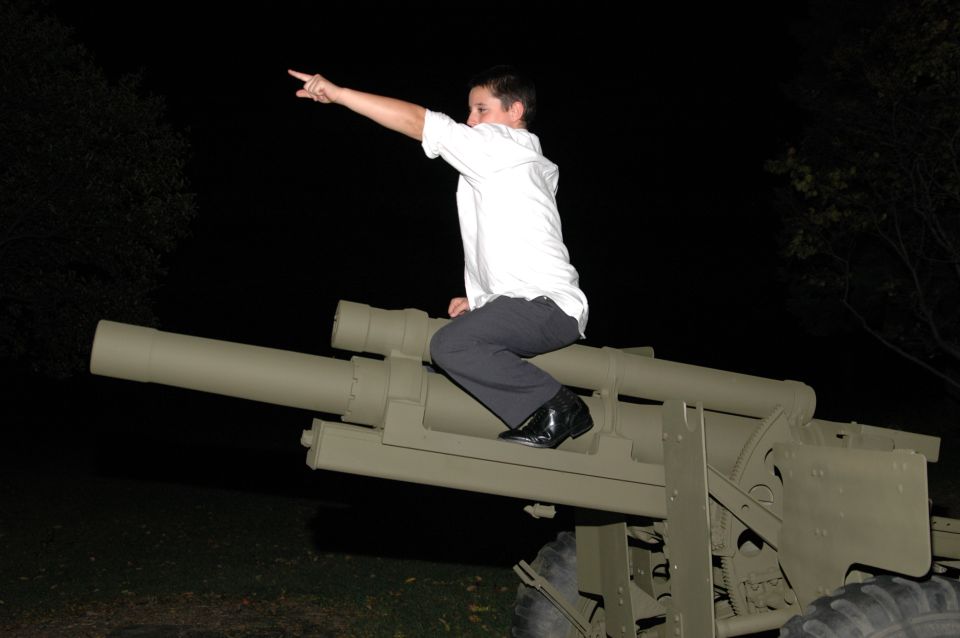 Person in white shirt and dark pants sits on a tan military howitzer, pointing left