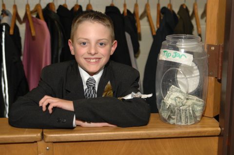 Boy in suit smiles next to a tip jar filled with money
