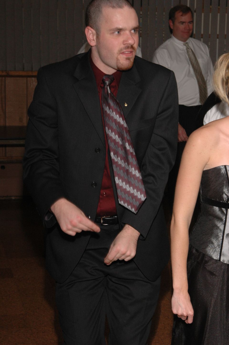 Man in a black suit, maroon shirt, and patterned tie gestures with his hands