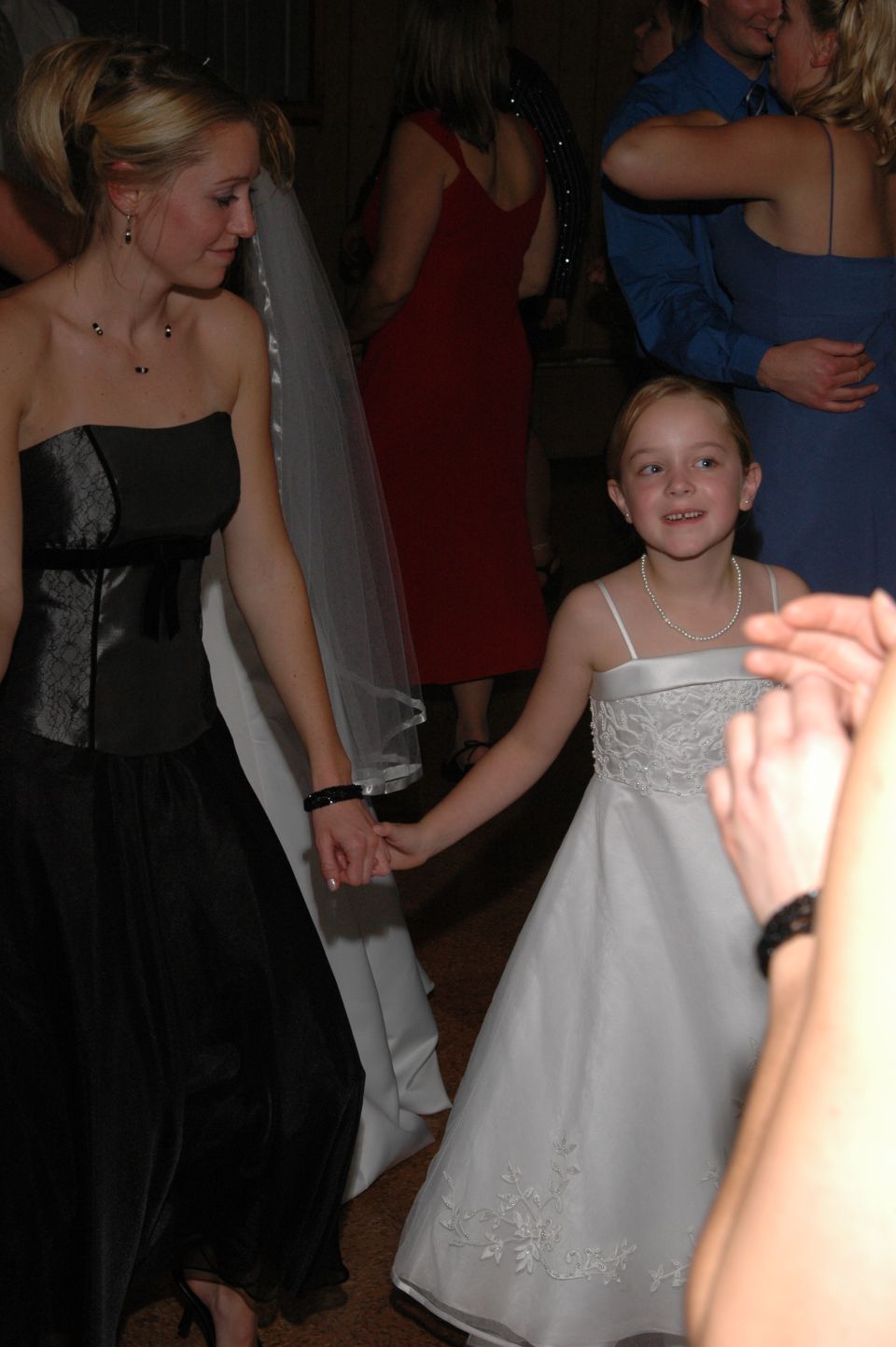 Young girl in white dress holds hands with woman in black dress at a wedding reception