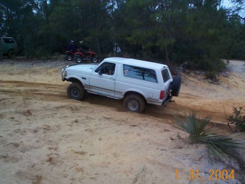1992 Ford Bronco - A white 1992 Ford Bronco navigates a sandy terrain, with other vehicles and trees visible.