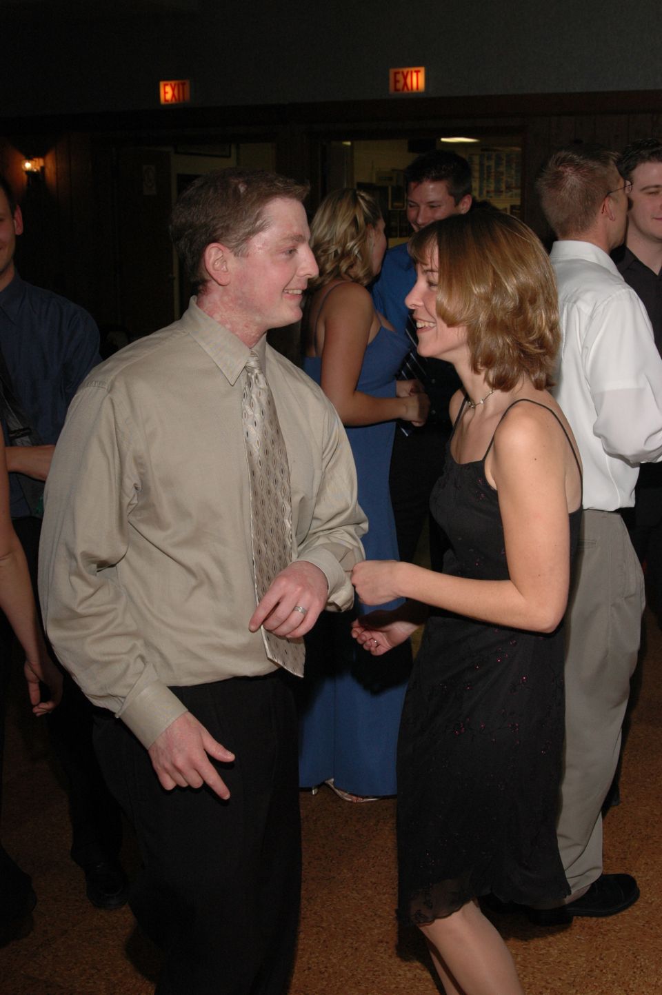 Man in tan shirt and tie dances with woman in black dress, other people in background