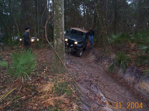 1992 Ford Bronco - A Jeep Wrangler navigates a muddy off-road trail in a wooded area.