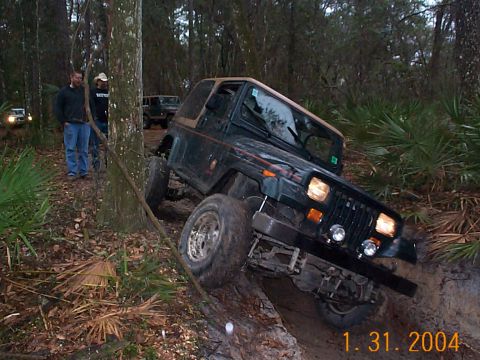 1992 Ford Bronco - A Jeep Wrangler is shown off-roading on a muddy, wooded trail with other vehicles present.