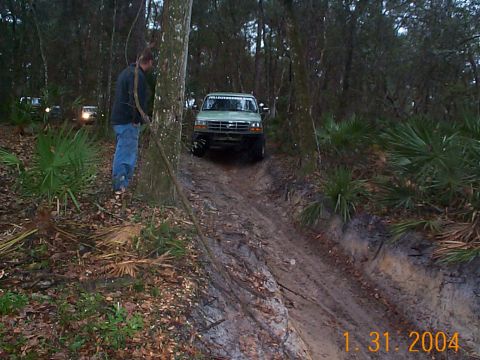 1992 Ford Bronco - A man watches as a Ford Bronco navigates a challenging off-road trail in a wooded environment.