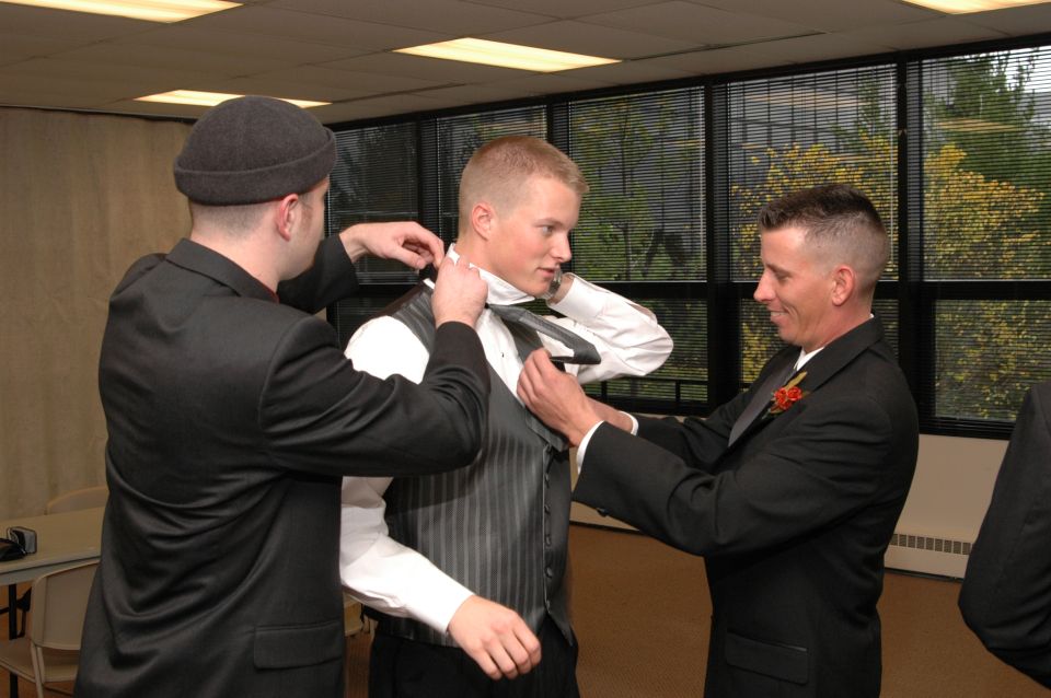 Groom in vest and white shirt having tie adjusted by two men in suits