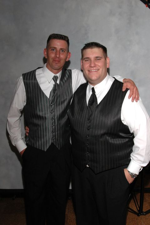 Two men in dark vests and ties stand with arms around each other