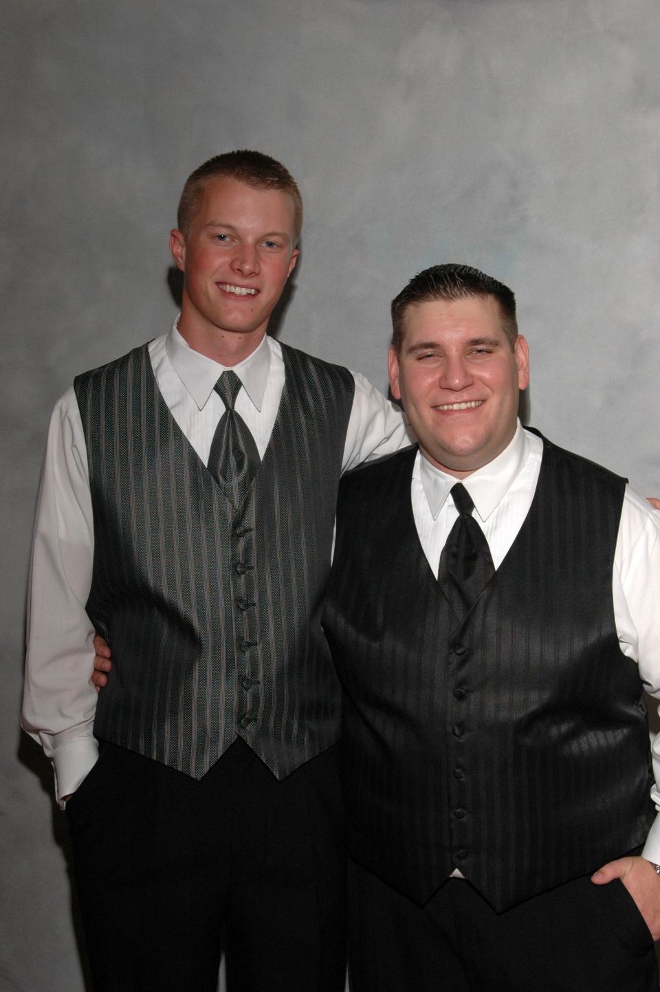 Two men in formal attire, one in a grey pinstripe vest and the other in a black pinstripe vest, pose for a photo