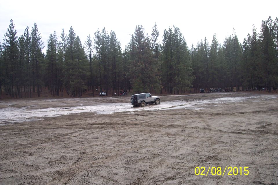 Silver Jeep Wrangler driving on a dirt track with trees in the background. Other vehicles are visible further back