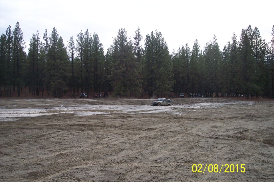 Silver 2005 Jeep Wrangler driving through a muddy area with trees in the background. Other vehicles are visible in the distance