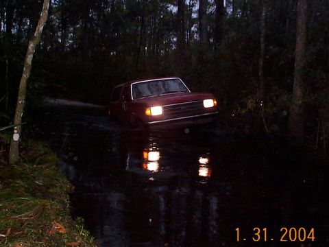 1992 Ford Bronco - A red SUV navigates through a flooded area surrounded by trees.