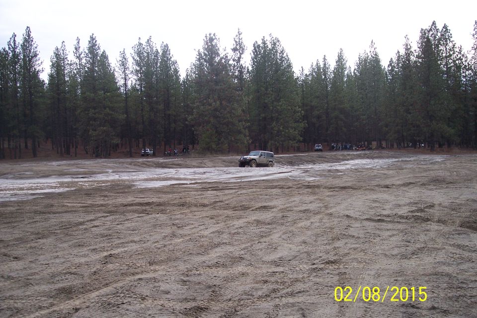 Jeep Wrangler driving through mud with trees in background. Other vehicles and people are visible further back
