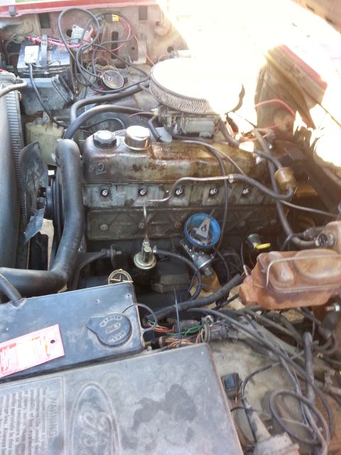 1981 Ford Bronco - Close-up view of a V8 engine bay, showing the carburetor, battery, and wiring. A warning label is present on a component.
