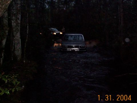 1992 Ford Bronco - Two SUVs navigate a muddy trail in a wooded area at dusk.