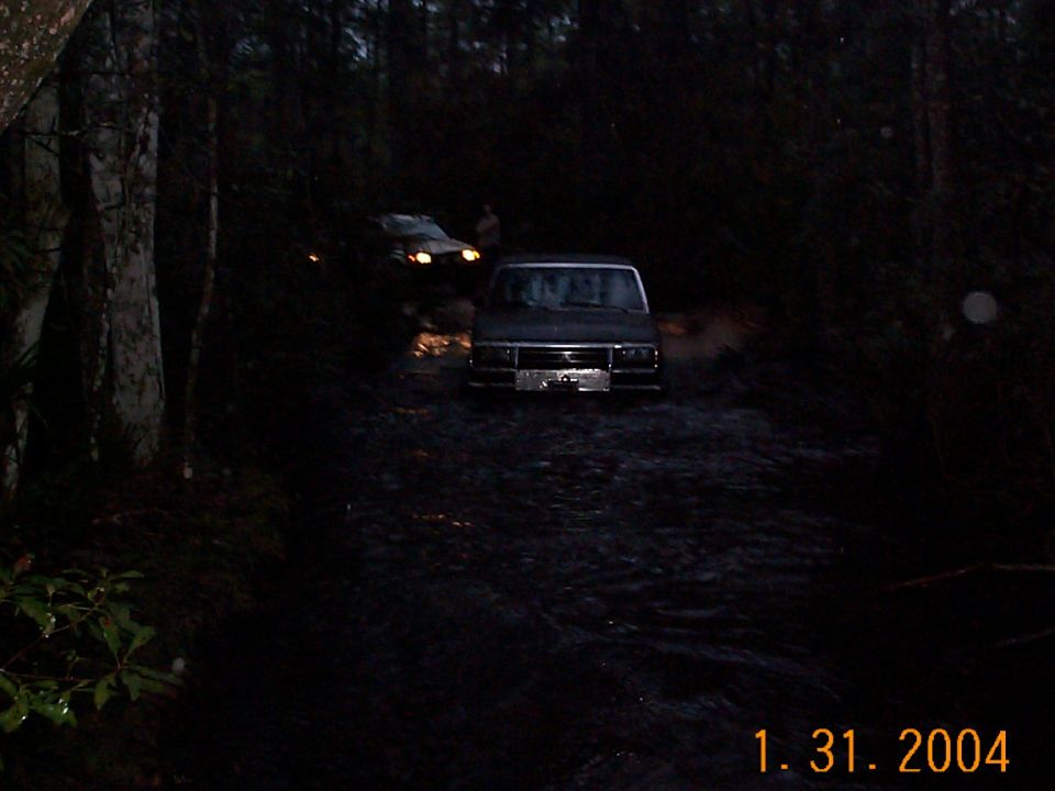 Dark SUV driving through mud with another vehicle behind it in a wooded area