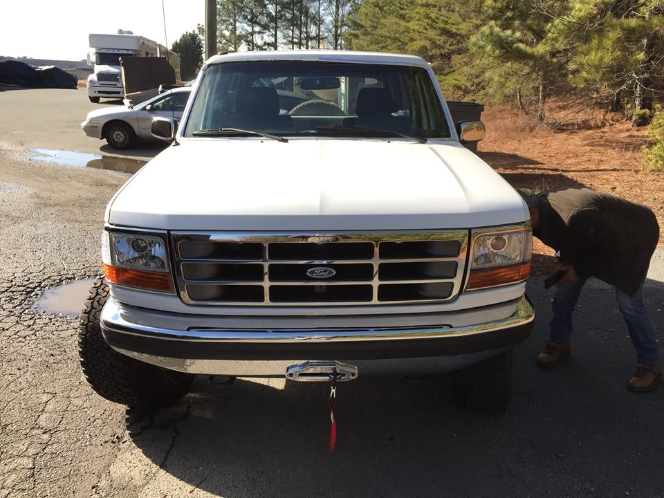 White Ford F-150 truck, front view, chrome grille, chrome bumper, aftermarket winch, person inspecting tire
