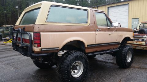 1988 Ford Bronco - A lifted, two-tone brown Ford Bronco sits outside a garage, with another vehicle visible in the background.