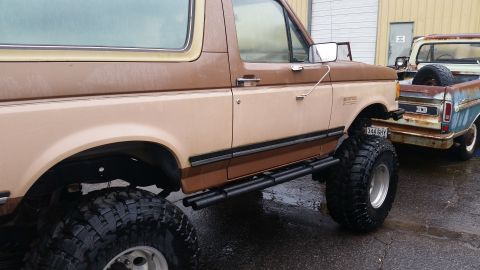 1988 Ford Bronco - A lifted Ford Bronco with a two-tone paint job sits beside an older, weathered pickup truck.