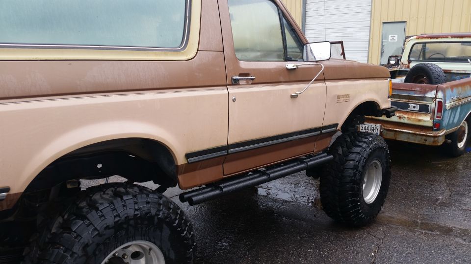 Brown and tan Ford Bronco with lifted suspension and large tires, parked next to a rusty pickup truck