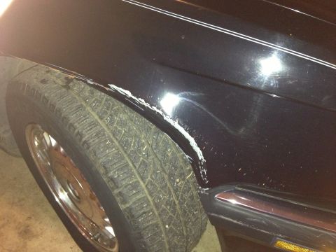 1988 BMW 7 Series - Close-up of damaged black car fender showing scraped paint and rust around the wheel well.