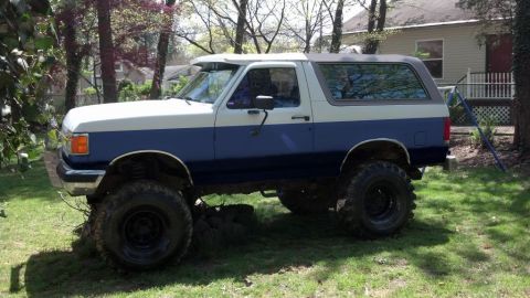 1994 Ford Bronco - Driver-side profile of a lifted two-tone blue and white bricknose Ford Bronco parked on grass in a wooded yard, with a grey removable hardt…