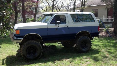 1994 Ford Bronco - Two-tone blue and white full-size Ford Bronco parked in profile on a grassy suburban yard, lifted on tall mud-terrain tires over black stee…