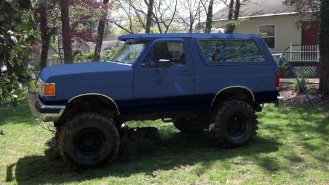1994 Ford Bronco - Blue lifted Ford Bronco full-size two-door SUV with oversized mud tires parked on a grassy yard near a house.