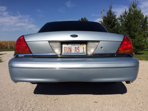 2003 Ford Crown Victoria - The rear of a light blue sedan is shown parked outdoors on a sunny day with a cornfield in the background.