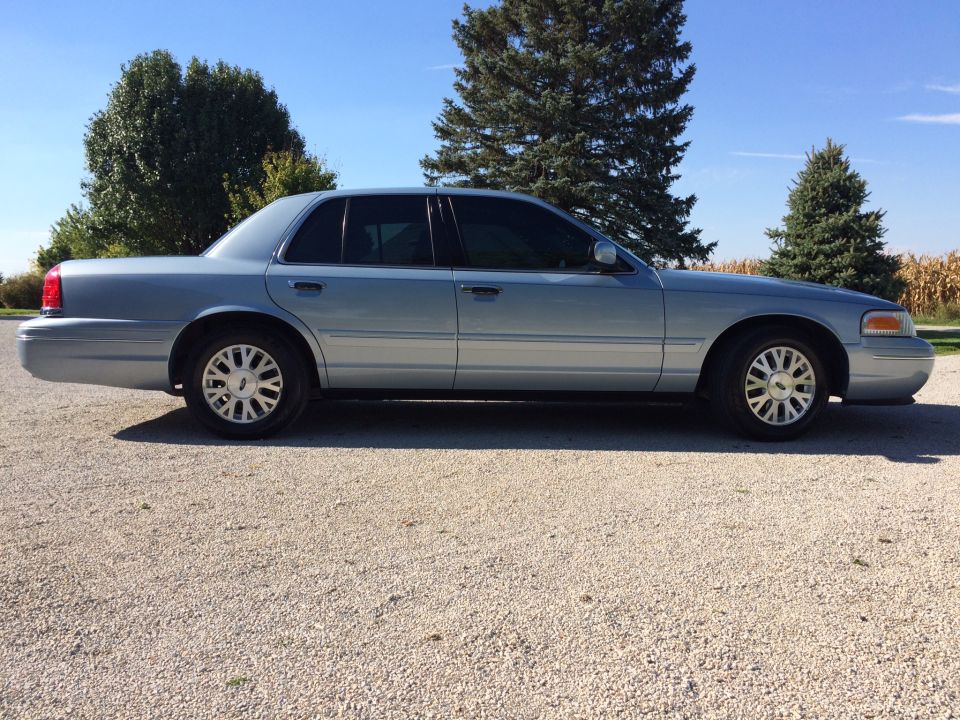 Side view of a 2003 Ford Crown Victoria parked on a gravel lot with trees and a cornfield in the background