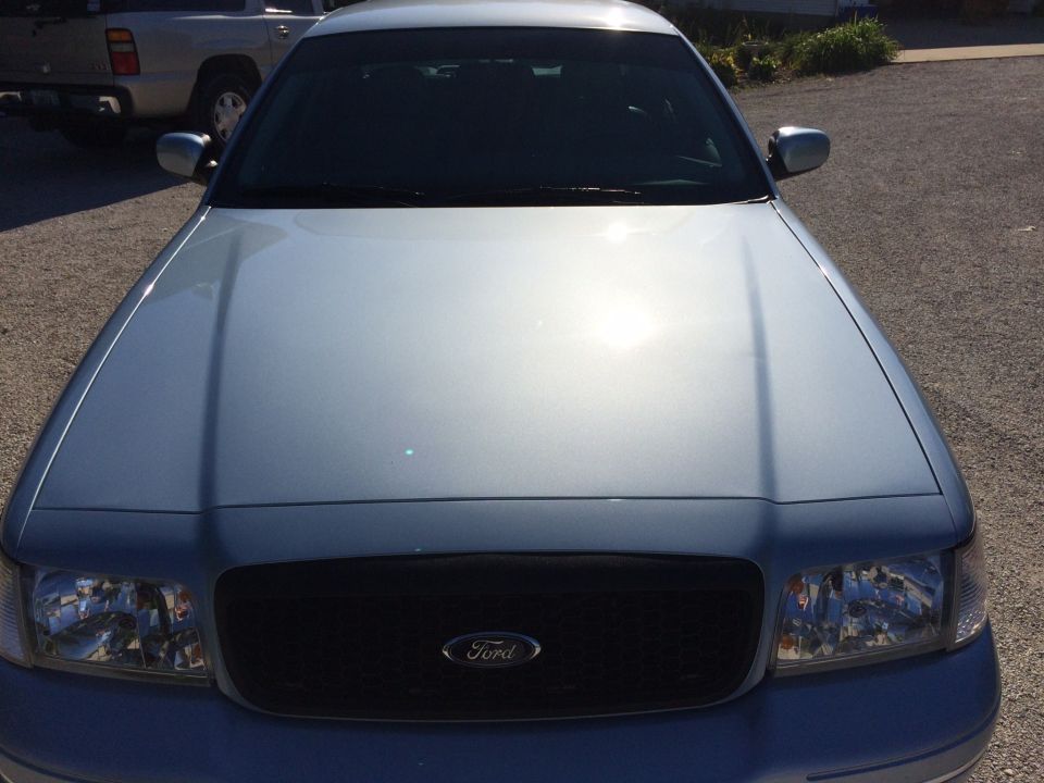 Front view of a light blue Ford Crown Victoria with a black grille. Another vehicle is parked behind it