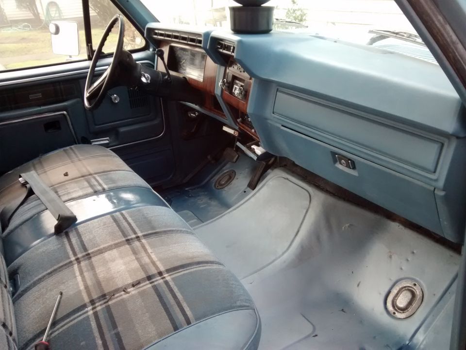 Interior of a blue 1980 Ford F-150 pickup truck, showing plaid bench seat, dashboard, and steering wheel