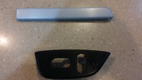 1995 Ford Bronco - Two interior trim pieces, one silver and one carbon fiber, are displayed on a speckled surface.