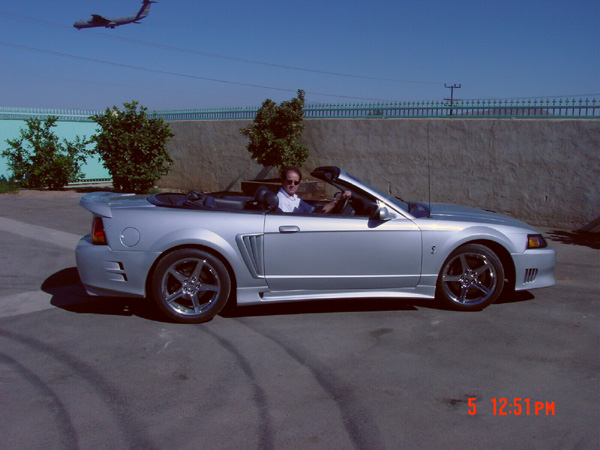 Silver Mustang convertible with body kit and chrome wheels, top down, driver seated, parked by a concrete wall