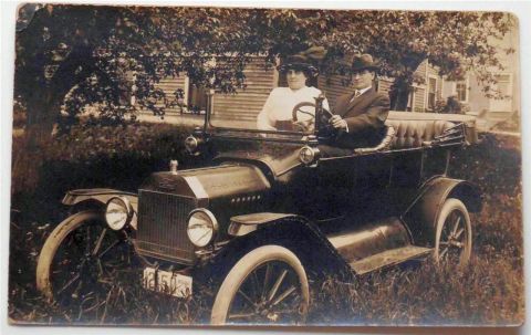 1915 Ford Model T - A man and woman pose in an open-top automobile, with a house and trees in the background.