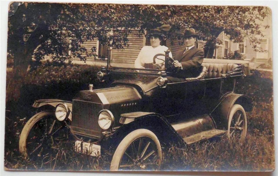 Couple in a touring car, possibly a Ford Model T, parked in front of a house