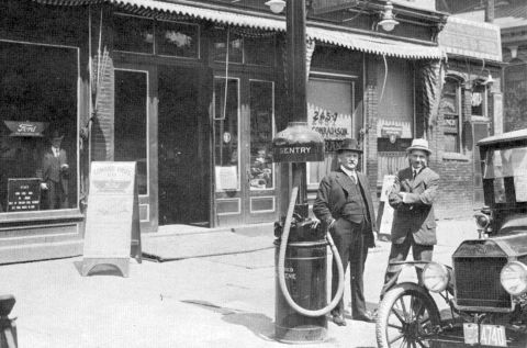 1915 Ford Model T - Two men stand by a vintage gas pump and a 1915 Ford Model T parked in front of a storefront.
